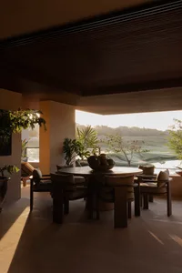 Round teak dining table with upholstered chairs on a covered patio overlooking a golf course at sunrise