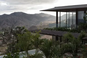 Modern mountain villa with large glass windows and a dark wooden terrace overlooking a vast tropical mountain valley under cloudy skies.