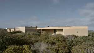 Modern single-story rammed earth coastal house with flat roof nestled among dense native coastal scrub under a blue sky.
