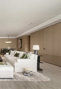 Modern minimalist living room featuring a large white linen sectional sofa with green velvet pillows, light oak wood panel walls, marble floor, and a contemporary pendant light in the background dining area.