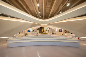 Modern exhibition hall interior featuring large curved concrete architectural elements, tiered pale yellow seating, and dramatic linear lighting along the ceiling.