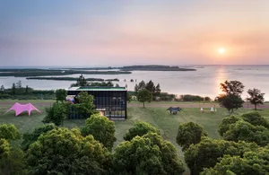 Modern black steel and glass pavilion with green roof situated on a grassy lakeside park at sunset, with pink and black shade sails in the foreground and a vast wetland landscape in the background.