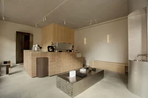 Modern minimalist cafe interior with a terracotta brick service counter, stainless steel coffee equipment, concrete floor, and textured metallic accents under warm lighting.
