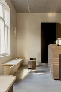 Minimalist cafe interior featuring a long light wood bench under a window, cylindrical wooden side tables with metallic accents, and a terracotta brick counter against a neutral beige wall.