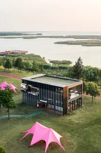 Aerial view of a modern two-story glass and metal commercial building with colorful facade art, situated on a green lawn next to a large lake with two bright pink tensile shade structures in the foreground.