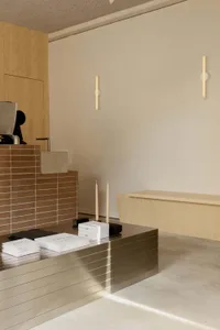 Modern minimalist reception desk made of horizontal stainless steel slats in the foreground, with a brown tiled counter and light wood cabinetry in the background, two tall candles and stacked brochures on the desk.