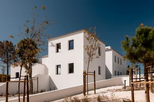Modern white stucco Mediterranean-style villas with flat roofs and small windows, surrounded by young trees and white boundary walls on sandy ground under a clear deep blue sky.