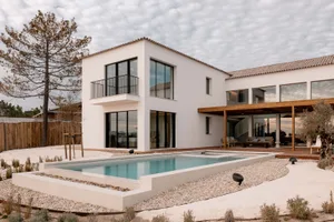 Modern white villa exterior with rectangular swimming pool, wooden pergola, outdoor lounge area, and gravel landscaping under a cloudy sky.