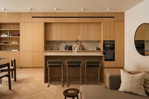 Modern open-plan kitchen featuring light oak cabinetry, a marble island with three dark green bar stools, herringbone wood flooring, and a neutral sofa in the foreground.