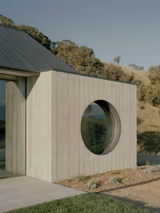 Modern concrete house exterior with a large circular window reflecting the surrounding hills and trees under a clear blue sky.