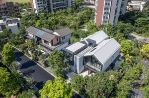 Aerial view of a contemporary luxury villa featuring extensive glass walls, corrugated metal cladding, and flat roofs, nestled among dense tropical trees in a suburban neighborhood.