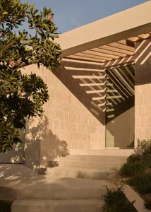 Modern minimalist house entrance featuring light beige travertine stone walls, wide concrete steps, wooden overhang, and strong diagonal shadows from a pergola, with a flowering tree on the left.