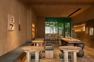 Modern wabi-sabi style restaurant interior with round solid wood tables, concrete walls, wooden stools with dark cushions, and large green-framed glass doors opening to a courtyard.