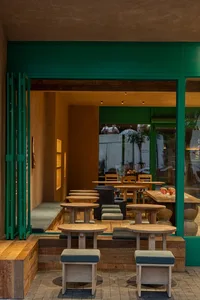 Modern cafe interior featuring round oak stools, built-in wooden banquettes with green cushions, and a bold emerald green entrance frame.
