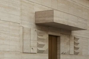Close-up of a luxurious travertine stone building entrance featuring geometric relief patterns, fluted columns, and a cantilevered overhang above a bronze door.