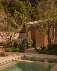 Modern teak wood cabana with vertical slats next to a natural swimming pool on a sunny hillside surrounded by native California landscaping and trees.