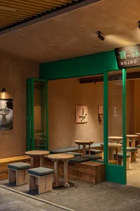 Modern Taiwanese restaurant interior with green metal frames, wooden round tables, built-in banquette seating with dark green cushions, textured beige walls, and warm lighting.