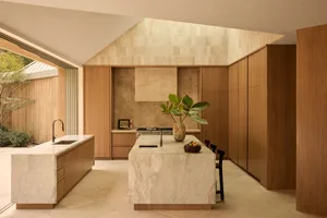 Modern kitchen featuring a large central island made of veined travertine, warm oak cabinetry, black metal chairs, and a tall skylight with textured stone walls.