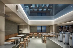 Modern minimalist café interior featuring raw concrete walls, stainless steel counters, large coffee grinders, and light oak wooden stools and tables under dramatic linear ceiling lighting.