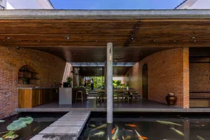 Modern tropical courtyard house featuring curved exposed brick walls, wooden ceiling, open kitchen, dining area, and a foreground koi pond with stepping stones.