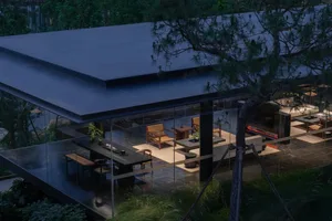 Night view of a modern glass villa with an open terrace featuring wooden furniture, a long dining table, and a fireplace, overlooking lush greenery with a prominent pine tree in the foreground.