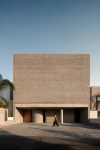 Front view of a modern brutalist brick building with a wooden door, cylindrical columns, and a woman walking across the paved courtyard under clear blue sky.