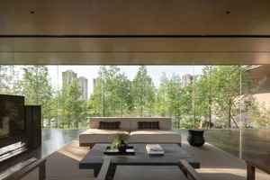 Modern minimalist living room featuring a low-profile cream sofa, dark stone coffee table, wood-paneled ceiling, and expansive floor-to-ceiling windows overlooking lush green trees and city buildings.