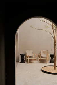 View through a dark arched doorway into a minimalist concrete courtyard featuring two wooden canvas sling chairs, black sculptural side tables, and a young tree planted in a circular gravel bed.