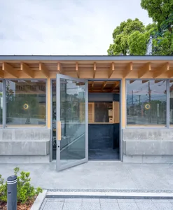 Modern minimalist building entrance featuring concrete base, light wood eaves, large glass windows, and an open grey pivot door revealing a dark interior with wooden storage.
