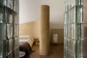 View through glass block walls into a minimalist bedroom featuring a large cylindrical concrete column, textured beige walls, wooden floor, and a bed with white and brown linen bedding.