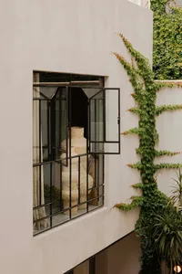 Modern minimalist exterior wall featuring a large black steel pivot window open to reveal tall woven rattan vases inside, with green ivy climbing the adjacent white concrete wall and potted plants below.