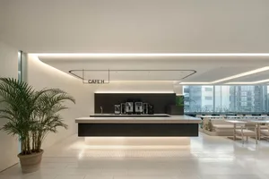 Modern minimalist cafe interior featuring a long dark wood service counter with white stone top, illuminated from below, a large potted palm tree in the foreground, and light wood seating area in the background.