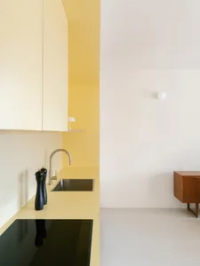 Modern minimalist kitchen featuring bright yellow walls, cream matte laminate cabinetry and countertops, stainless steel sink and faucet, black induction cooktop, and a wooden side cabinet against a white wall.