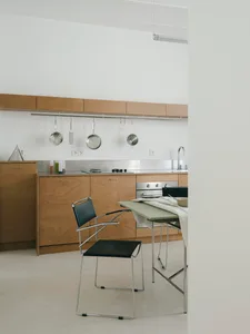 Minimalist kitchen interior featuring light plywood cabinetry, stainless steel backsplash, hanging cookware, and a black leather cantilever chair with chrome frame next to a sage green dining table.