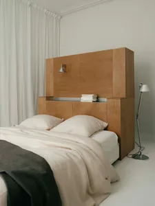 Modern minimalist bedroom with a large oak wood storage headboard, white linen bedding, dark grey throw blanket, and a chrome floor lamp against white walls and curtains.