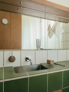Close-up of a modern bathroom vanity featuring glossy green ceramic tiles, a stainless steel wall-mounted faucet, oak mirror cabinet, and toiletries on the counter.