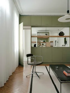 Modern living room featuring sage green built-in cabinetry, a black leather and chrome swivel armchair, a black marble coffee table, and a white area rug with black stripes on herringbone parquet flooring.