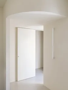 Minimalist white hallway featuring a curved archway, flush white door, and vertical wall sconce in a serene monochromatic palette.
