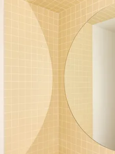 Close-up view of a bathroom corner featuring butter yellow square ceramic tiles, white grout, and a large round mirror with a thin gold frame.