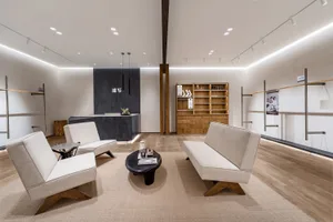 Modern minimalist furniture showroom waiting area with white linen sofas, oak bases, black coffee table, wooden shelving unit, and dark accent wall under bright linear lighting.