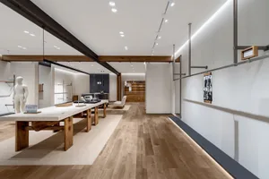 Long view down a minimalist furniture showroom with large oak and concrete display tables, light wood flooring, recessed ceiling lights, and wooden beams.
