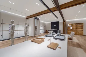 Modern minimalist furniture showroom interior with oak wood elements, white concrete display tables, wooden beams, and mannequin displays under warm lighting.