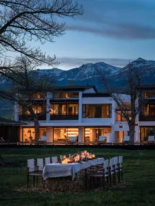 Elegant outdoor dining setup using hay bales as seating base with white tablecloth and candlelight, in front of a modern white farmhouse with illuminated interiors and snow-capped mountains in the background at twilight.