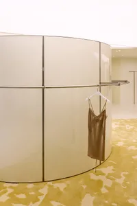 Curved high-gloss cream lacquered wall in a boutique with a brown silk slip dress hanging on a white hanger, set on a textured chartreuse carpet.