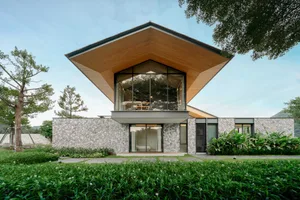 Modern two-story house with a dramatic floating wooden roof, large glass windows, and stone-clad lower level, surrounded by lush tropical garden.