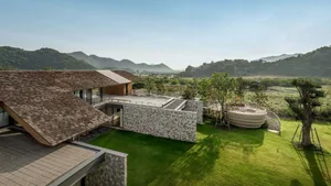 Aerial view of a modern luxury villa featuring stone walls, wooden shingle roofs, and a circular terrace overlooking green mountains and fields in Taiwan.