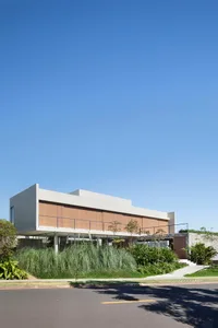Modern two-story concrete and wood house with elevated structure, horizontal timber facade, large glass windows, and lush tropical landscaping with tall ornamental grasses under a clear blue sky.