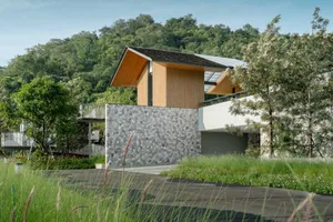 Modern mountain house featuring a lower level of stacked natural stone, upper floors clad in vertical timber slats, large glass openings, and dramatic roof overhangs, set against a green forested hillside with wild grasses in the foreground.