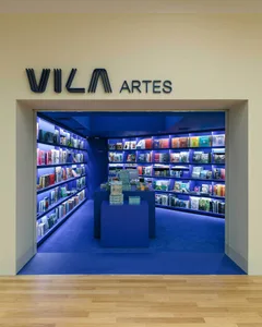 View into a contemporary bookstore with intense blue lighting, floor-to-ceiling bookshelves, central display islands, and a large blue sign reading VILA ARTES above the entrance.