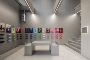 Modern minimalist sneaker retail store interior with raw concrete walls, linear LED lighting, glass display table, and colorful sneaker wall display arranged in a grid.